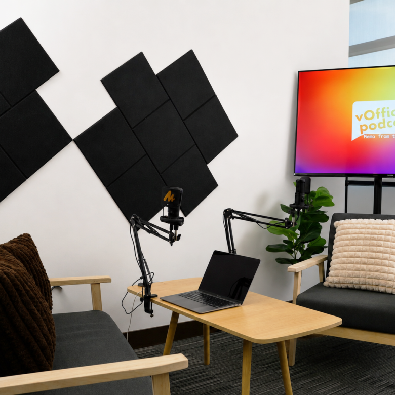 Podcast recording setup with two gray chairs, a wooden table, laptop, and dual boom microphones in front of a white wall with black acoustic panels; screen shows 'vOffice podcast'.