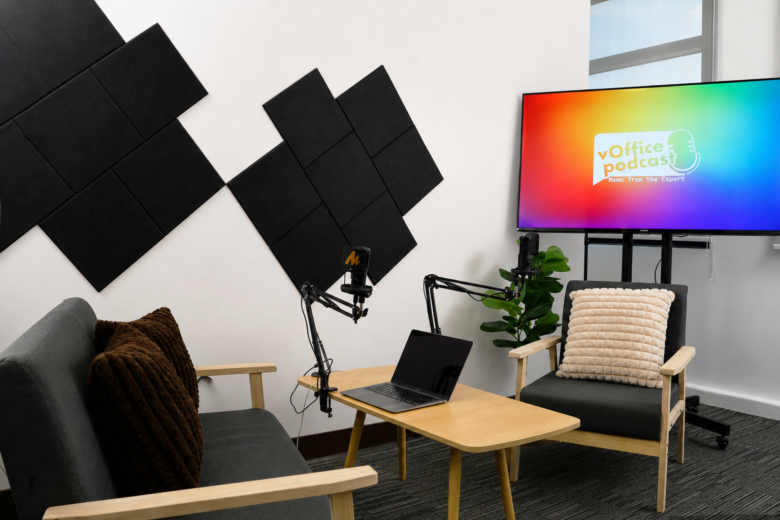 Podcast recording setup with two gray chairs, a wooden table, laptop, and dual boom microphones in front of a white wall with black acoustic panels; screen shows 'vOffice podcast'.